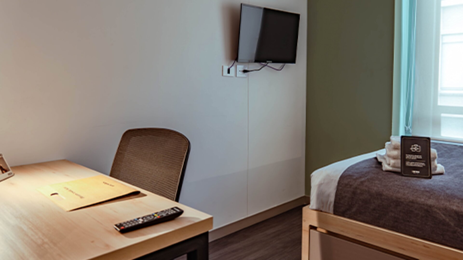 Compact guest room with a single bed, dark bed runner, wooden desk, chair and wall-mounted television at UMA Suites Chía.
