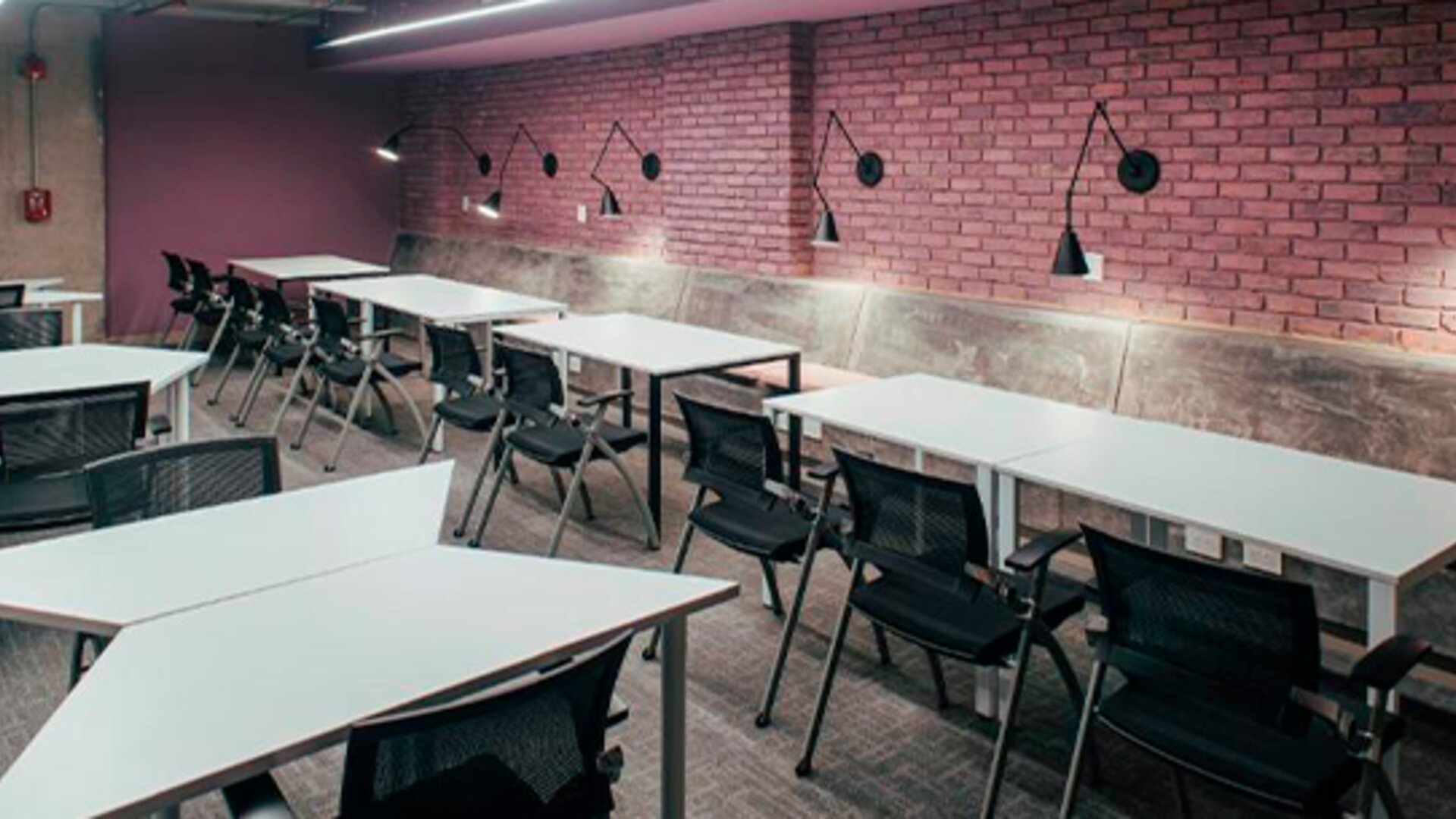 Communal study area with white tables, black chairs, exposed brick walls and wall-mounted lamps at UMA Suites Chía.