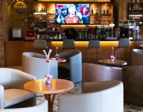 Hotel bar and lounge with upholstered armchairs, round coffee tables, a wooden bar counter and a television at Hotel Habitel Select.