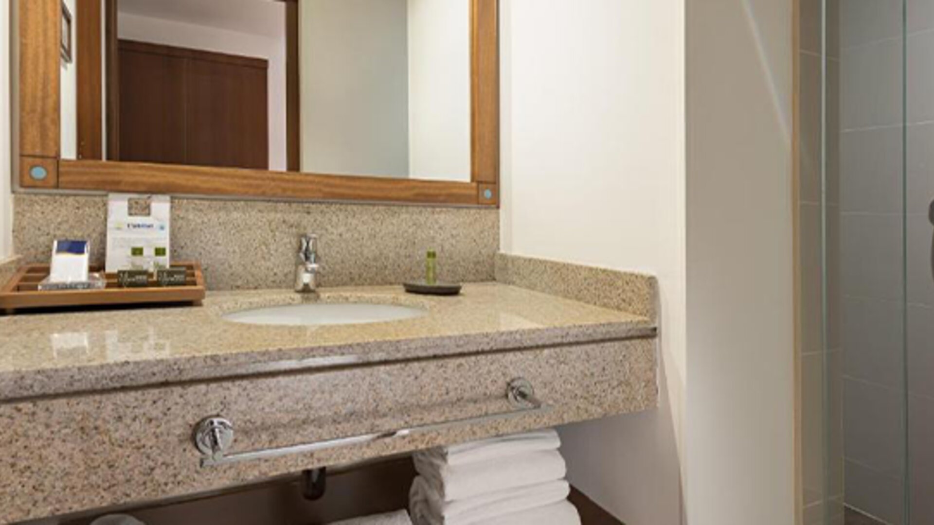 Guest bathroom with a granite vanity, inset washbasin, wooden-framed mirror, folded towels and glass-enclosed shower at Hotel Habitel Select.