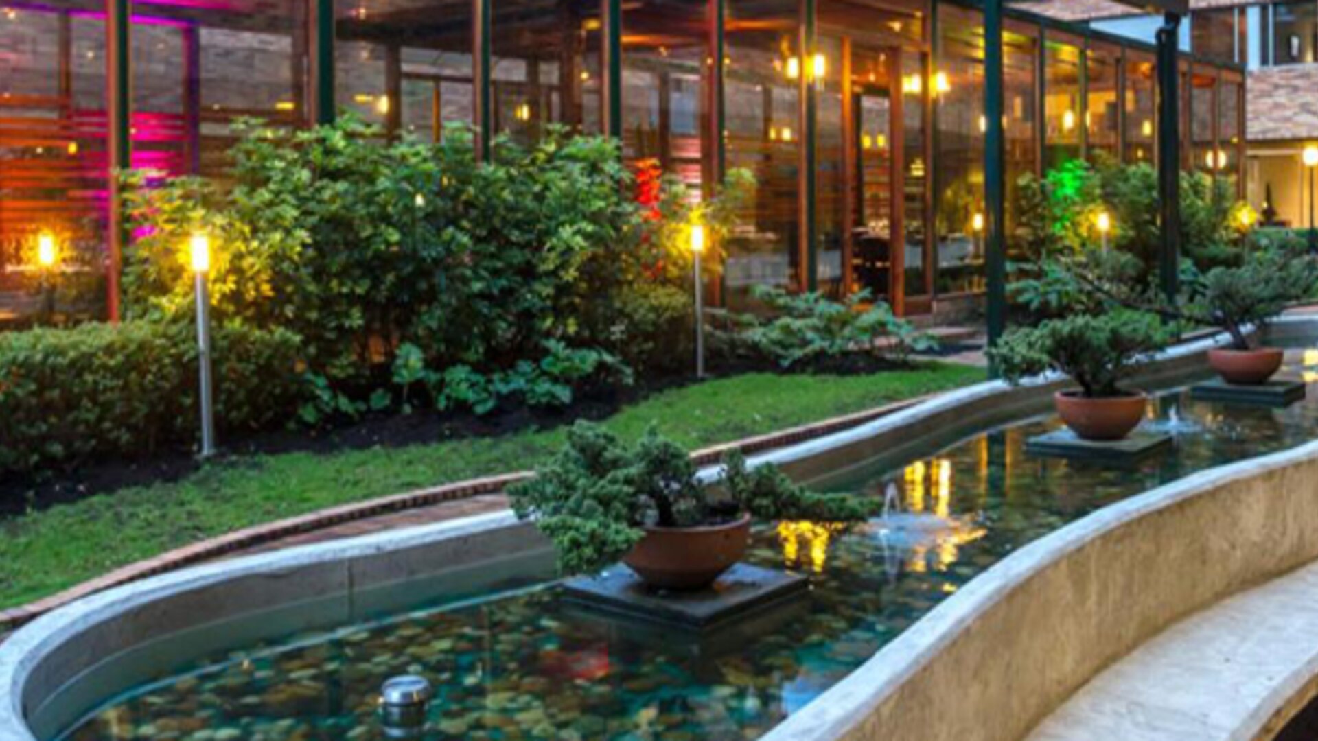 Curved ornamental pond with water fountains, planted greenery and outdoor lighting beside the hotel at Hotel Habitel Select.