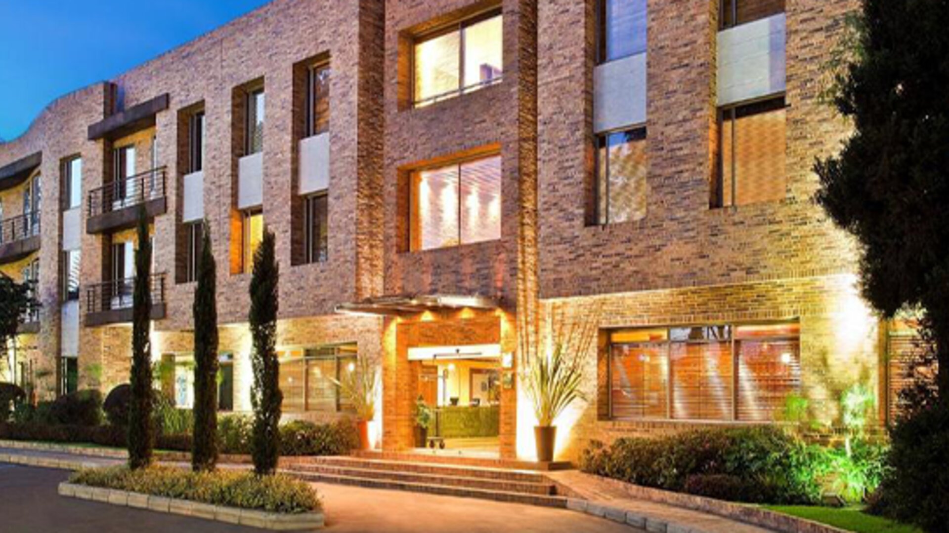 Brick hotel exterior illuminated at dusk, with landscaped gardens, tall windows and a warmly lit entrance at Hotel Habitel Select.