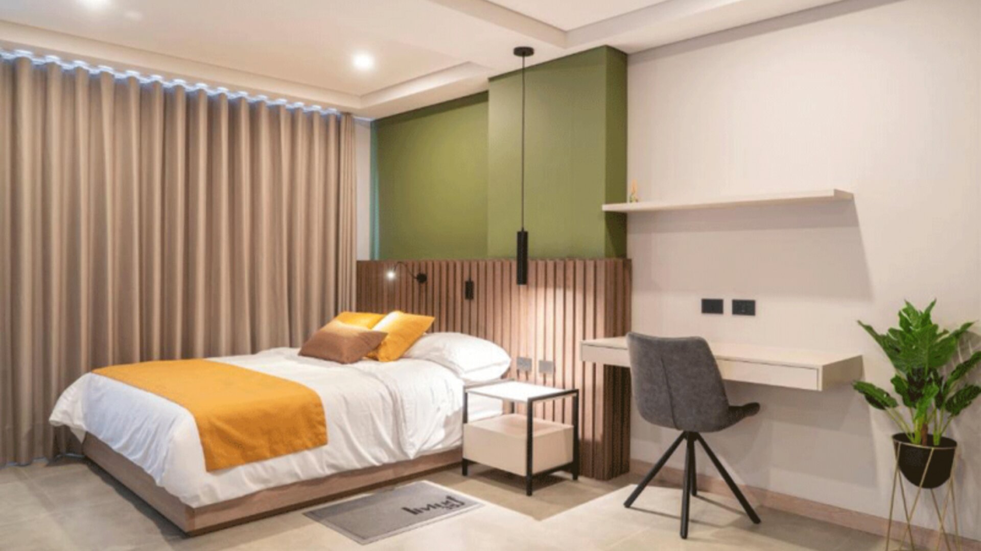 Guest bedroom with a double bed, mustard and brown cushions, green accent wall, wooden slat headboard, desk, and potted plant at Hotel Living 35 Suites.