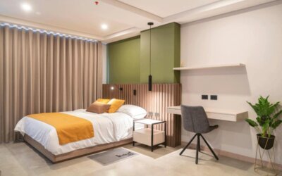 Guest bedroom with a double bed, mustard and brown cushions, green accent wall, wooden slat headboard, desk, and potted plant at Hotel Living 35 Suites.