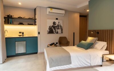 Guest suite with a double bed, teal kitchenette cabinetry, wooden headboard, wall-mounted television, and compact seating area at Hotel Living 35 Suites.