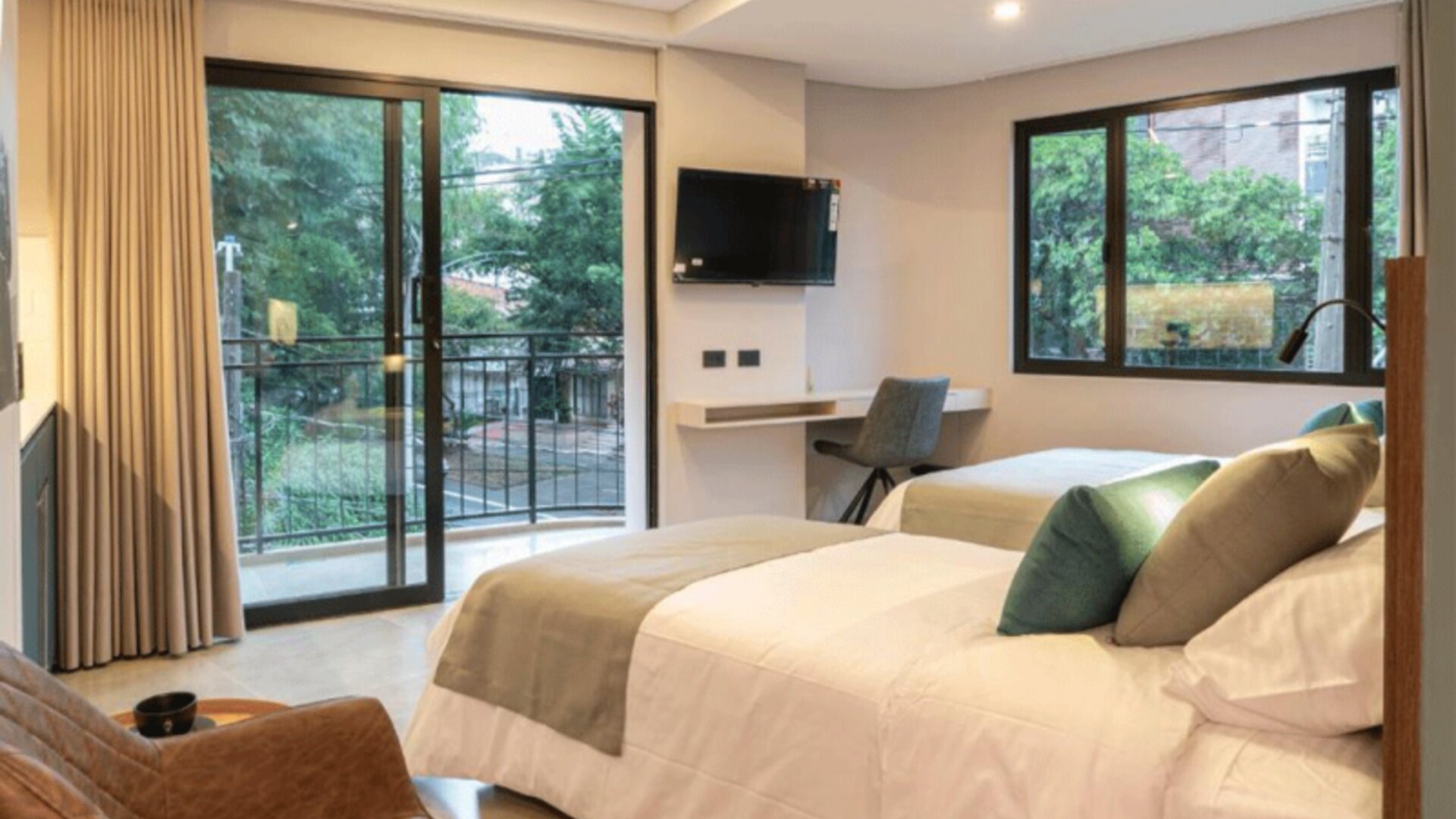 Guest bedroom with two beds, neutral bedding, teal and green accent cushions, a wall-mounted television, desk, and balcony doors at Hotel Living 35 Suites.