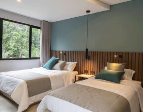 Guest room with two beds dressed in white linens, teal accent cushions, a wooden slat headboard, and large windows at Hotel Living 35 Suites.
