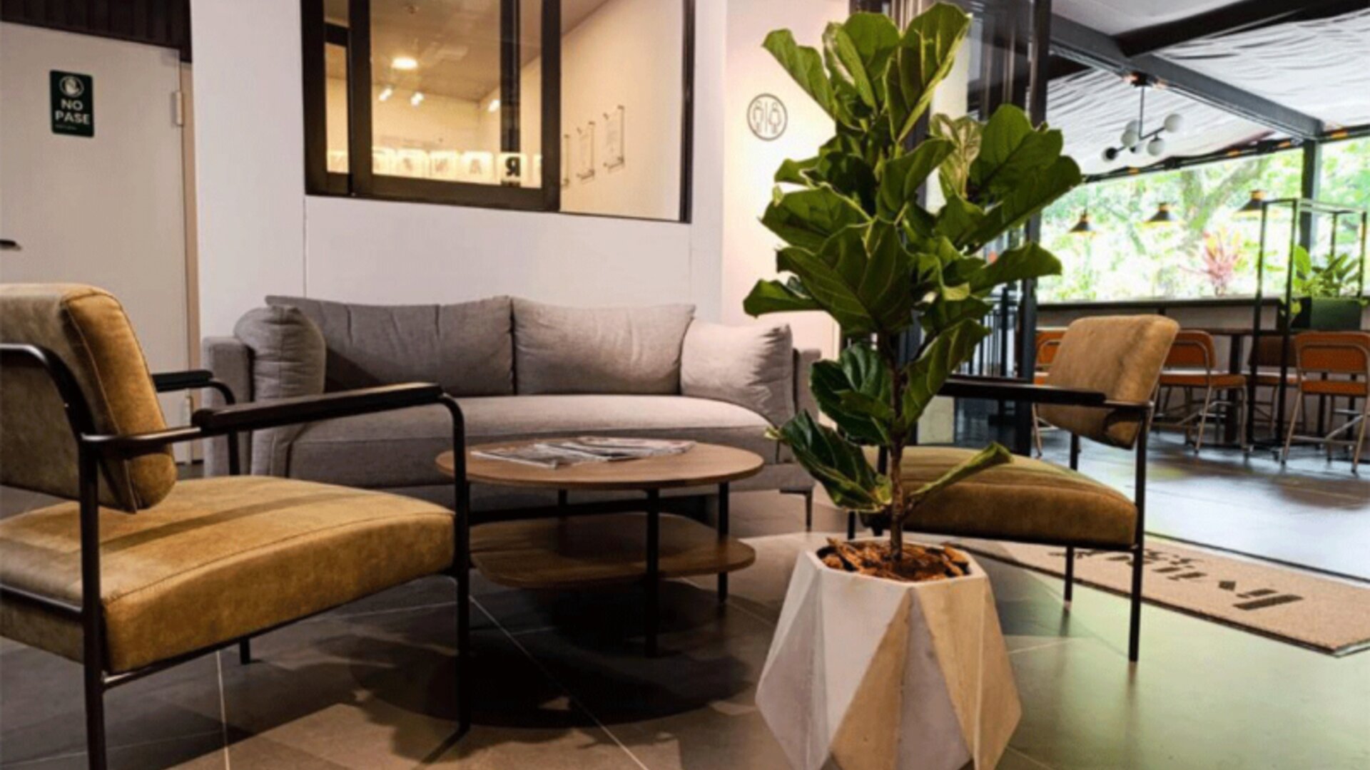Lounge area with a grey sofa, mustard-coloured armchairs, round coffee table, large potted plant, and adjoining café seating at Hotel Living 35 Suites.