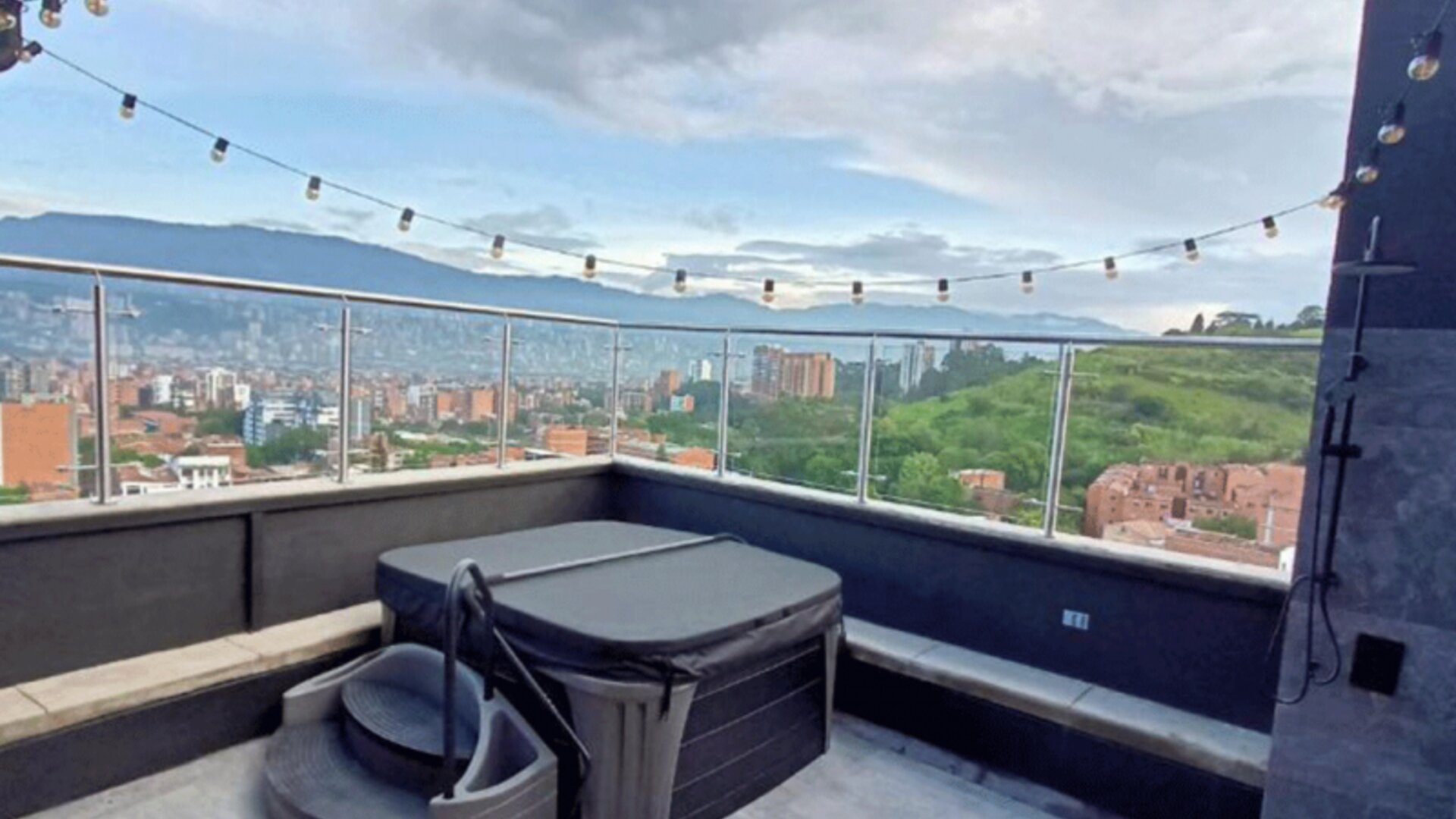 Rooftop terrace with a hot tub, built-in seating, glass railings, string lights, and panoramic city and mountain views at Hotel Living 35 Suites.