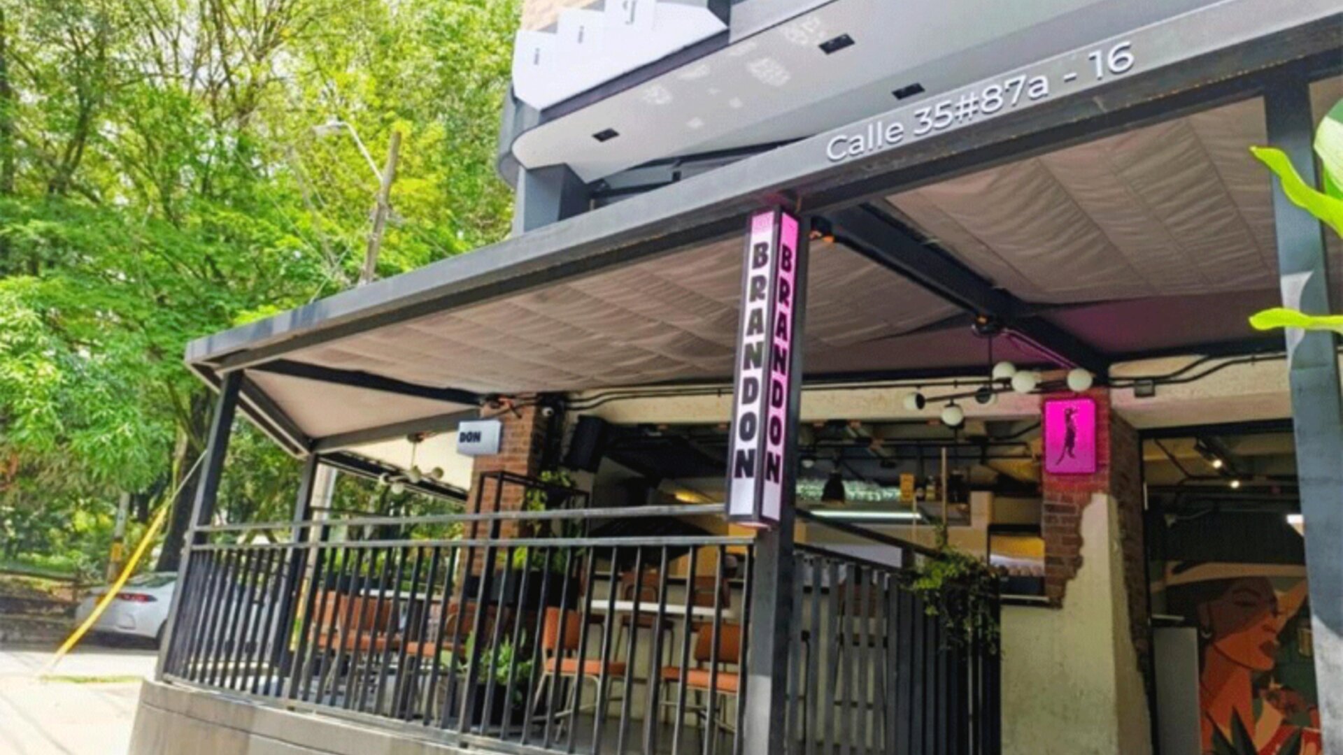 Street-level entrance with a covered terrace, black railings, vertical signage, and surrounding greenery at Hotel Living 35 Suites.