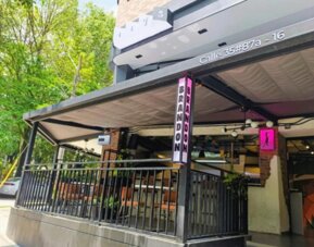 Street-level entrance with a covered terrace, black railings, vertical signage, and surrounding greenery at Hotel Living 35 Suites.