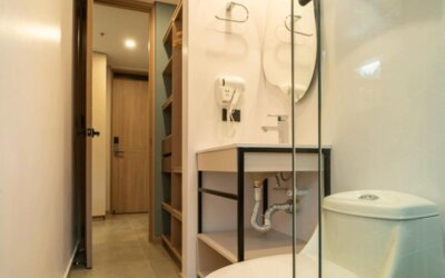 Compact bathroom with a white toilet, open-shelf vanity, wall-mounted hairdryer, glass shower partition, and beige tiled flooring at Hotel Living 35 Suites.