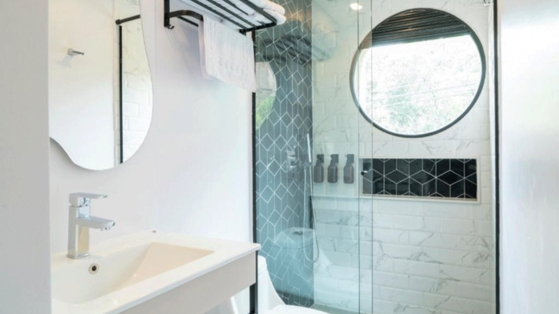 Modern bathroom with a white vanity, curved mirror, glass-enclosed shower, white and grey patterned tiles, and a round window at Hotel Living 35 Suites.