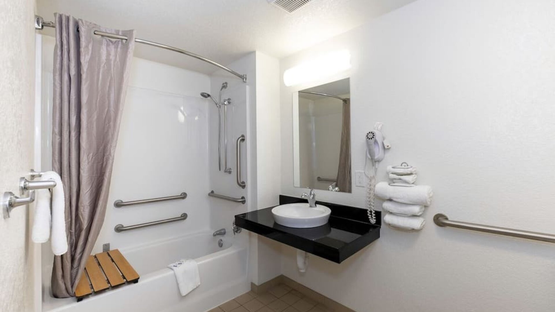 Accessible bathroom with a bathtub, shower curtain, grab bars, wall-mounted mirror, black countertop, and white vessel sink at American Inn Carlisle.