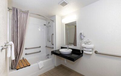Accessible bathroom with a bathtub, shower curtain, grab bars, wall-mounted mirror, black countertop, and white vessel sink at American Inn Carlisle.