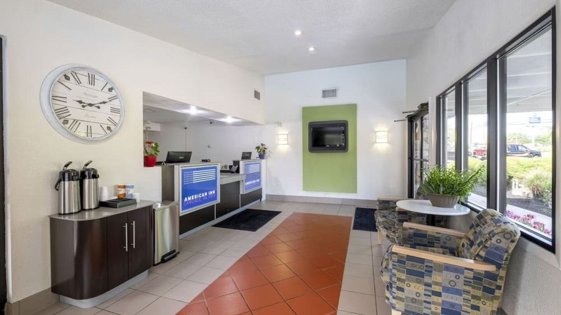 Reception and lobby area with tiled flooring, a front desk, a large wall clock, beverage station, seating, and windows at American Inn Carlisle.