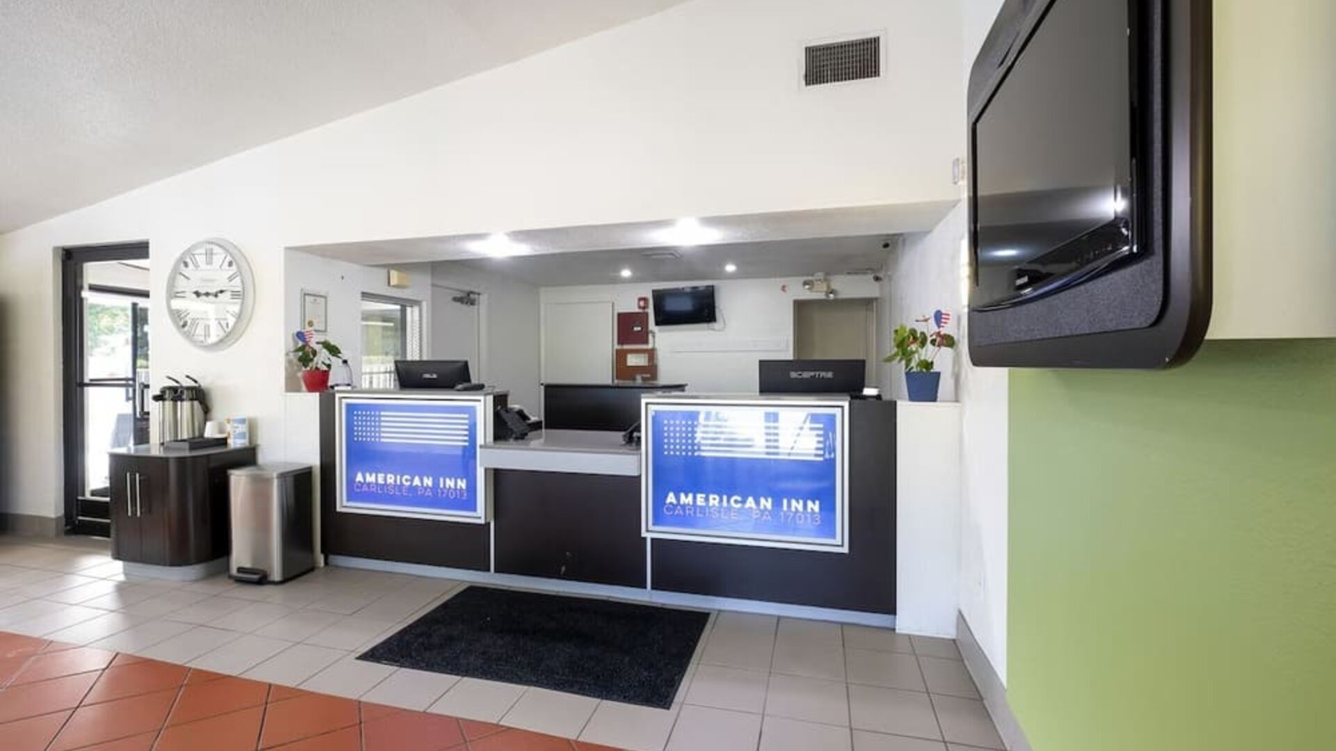 Reception desk with American Inn Carlisle signage, computers, a wall-mounted television, and a tiled lobby floor.