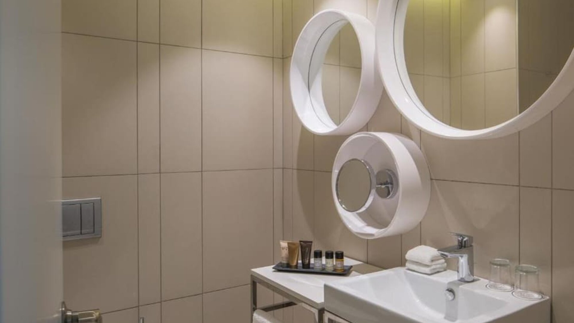 Contemporary bathroom with beige tiled walls, a white vanity sink, circular mirrors, toiletries, and neatly folded towels at Prima Millennium.