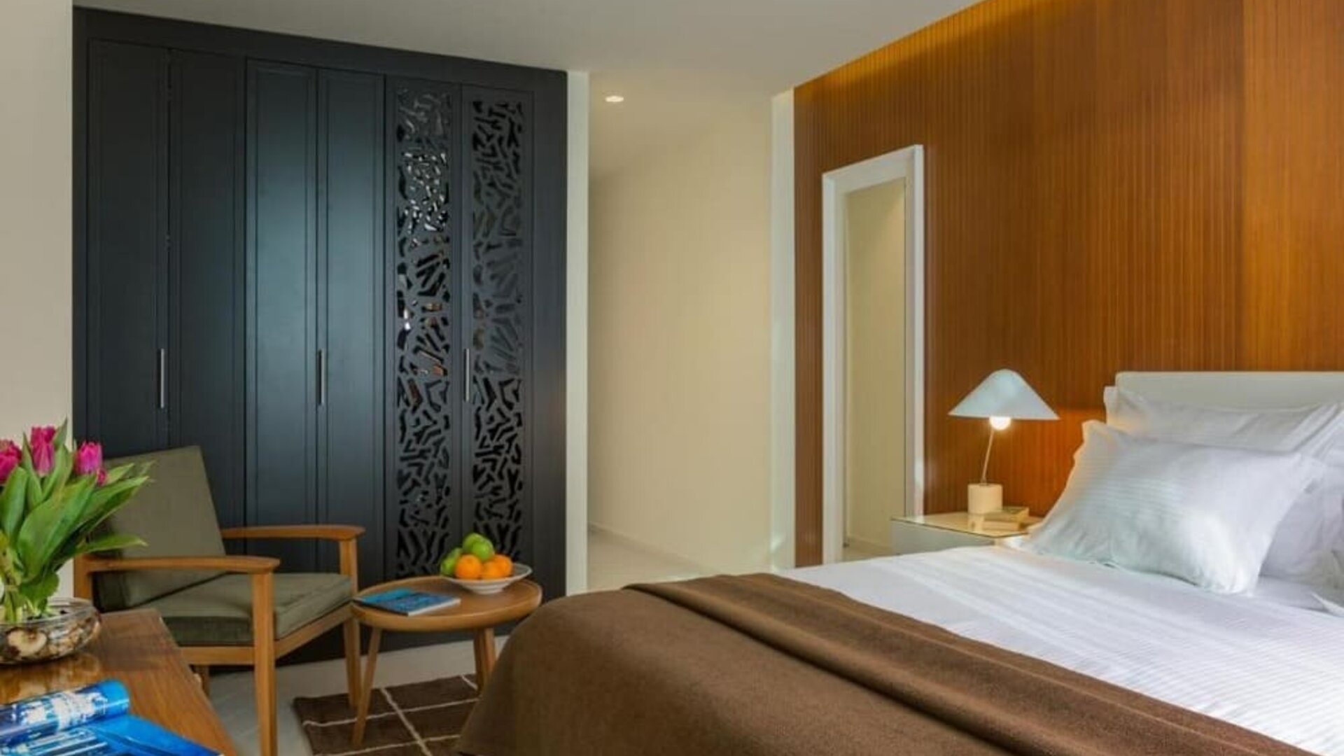 Guest room with a king-size bed, white bedding, a brown throw, wood-panelled accent wall, bedside lamps, wardrobe, and seating area at Prima Millennium.