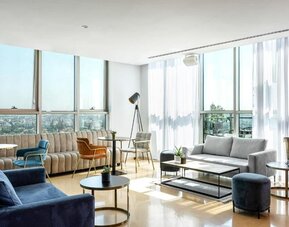 Spacious hotel suite lounge with floor-to-ceiling windows, city views, blue and neutral sofas, accent chairs, and contemporary coffee tables at Prima Millennium.