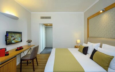 Guest bedroom with a king-size bed, white bedding, a gold bed runner, bedside tables, a wall-mounted television, and a desk with a chair at Prima Park Hotel.