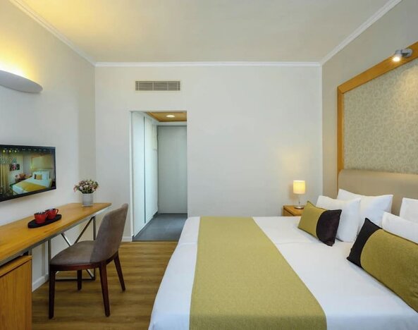 Guest bedroom with a king-size bed, white bedding, a gold bed runner, bedside tables, a wall-mounted television, and a desk with a chair at Prima Park Hotel.