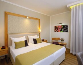Guest room with a king-size bed, white bedding, gold and dark accent cushions, a wooden headboard frame, and a small seating area beside the window at Prima Park Hotel.