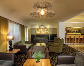 Hotel lounge with upholstered armchairs and sofas, wooden coffee tables, patterned carpeting, decorative pendant lighting, and an adjoining bar at Prima Park Hotel.