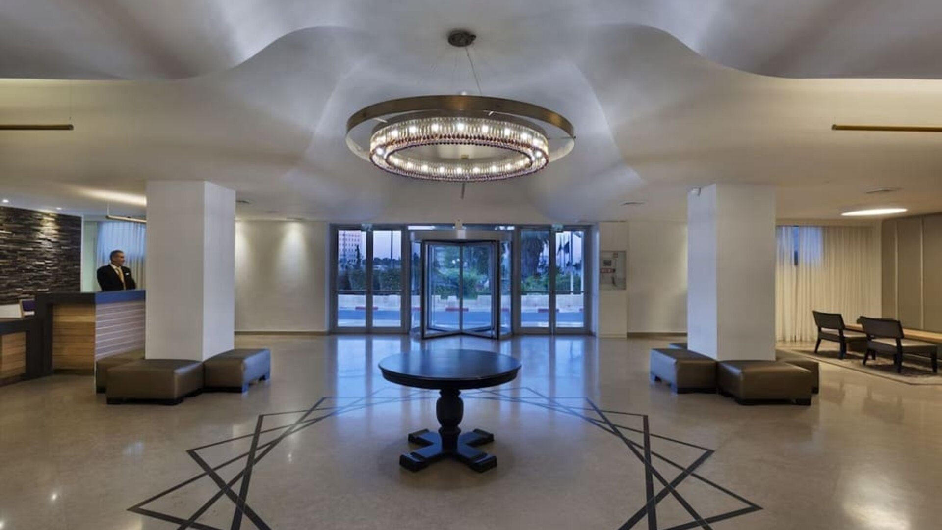 Hotel reception lobby with a front desk, polished flooring, a circular chandelier, seating areas, and glass entrance doors at Prima Park Hotel.