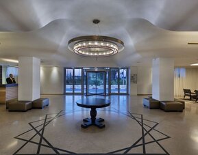 Hotel reception lobby with a front desk, polished flooring, a circular chandelier, seating areas, and glass entrance doors at Prima Park Hotel.