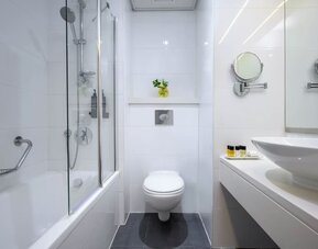 Modern bathroom with a bathtub and glass shower screen, wall-mounted toilet, white vanity basin, magnifying mirrors, and toiletries at Prima Park Hotel.