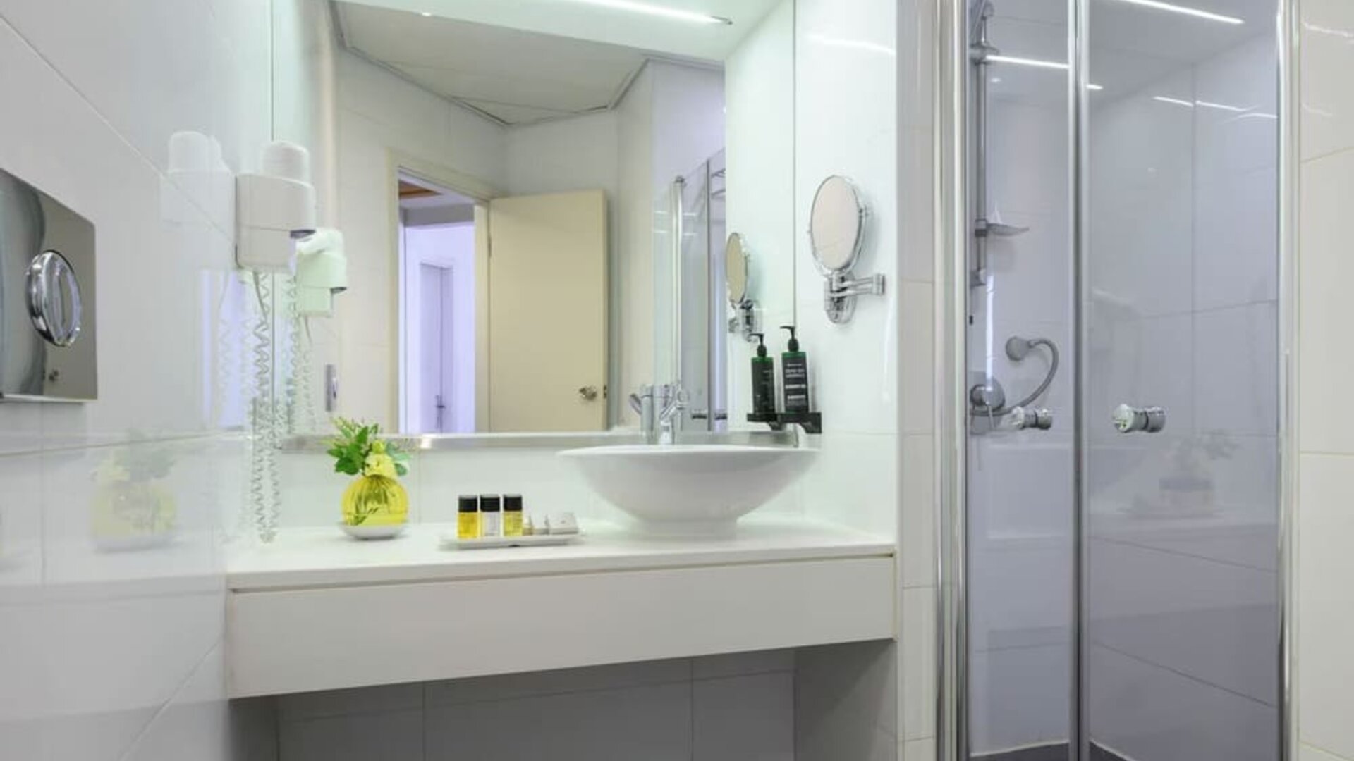 Bright hotel bathroom featuring a white vanity with a vessel sink, large mirror, glass-enclosed shower, wall-mounted hairdryer, and toiletries at Prima Park Hotel.