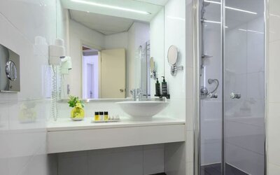 Bright hotel bathroom featuring a white vanity with a vessel sink, large mirror, glass-enclosed shower, wall-mounted hairdryer, and toiletries at Prima Park Hotel.