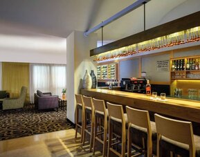 Hotel bar with a long wooden counter, bar stools, illuminated bottle shelves, glassware, and an adjoining lounge seating area at Prima Park Hotel.