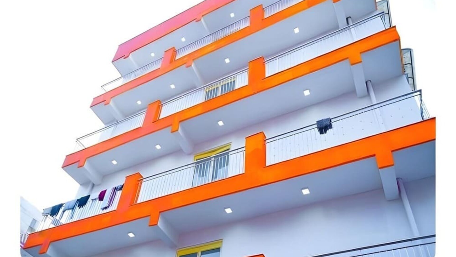 Exterior view of the multi-storey hotel building featuring white walls, bright orange balcony accents, metal railings, and balconies overlooking the surrounding area.