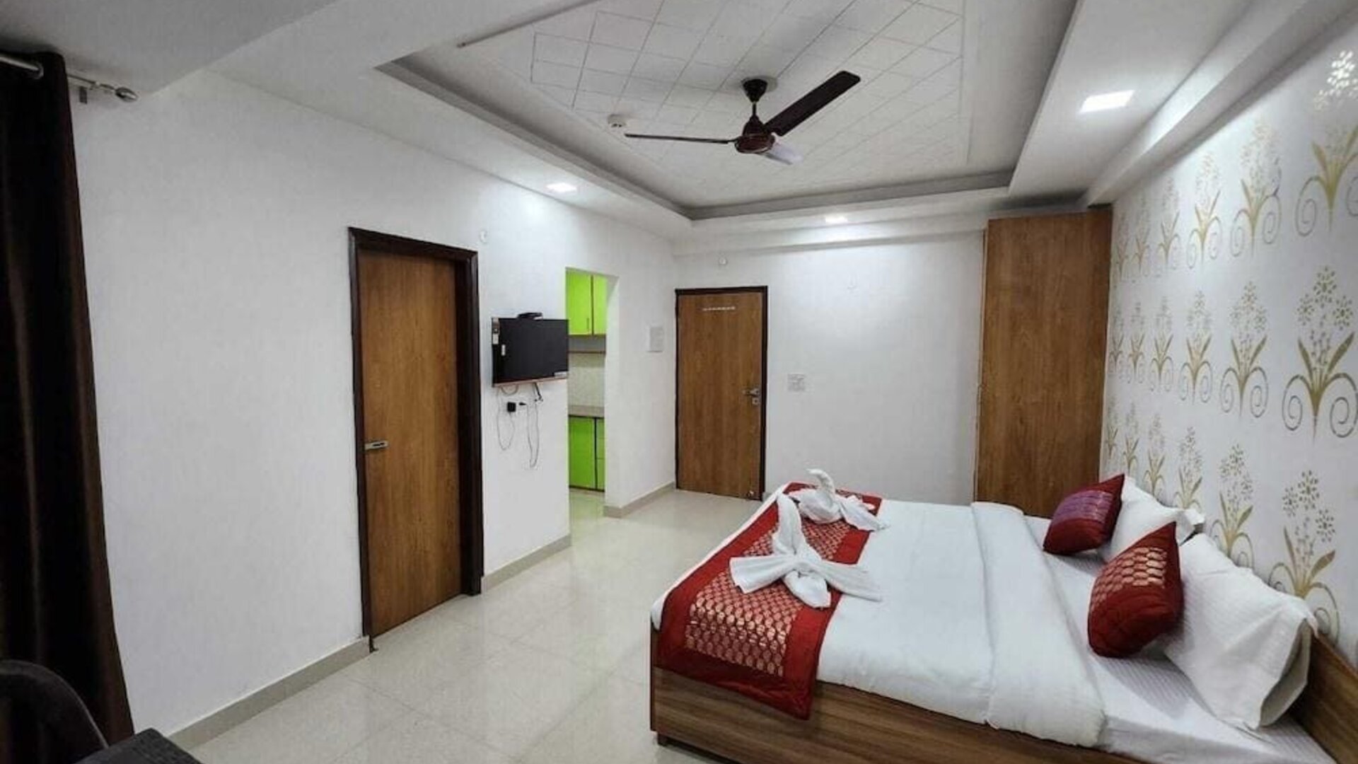 Spacious guest room with a double bed dressed in white bedding and red accent cushions, a red bed runner, decorative feature wall, wooden wardrobes, wall-mounted television, and tiled flooring at Hotel Urban Abode.
