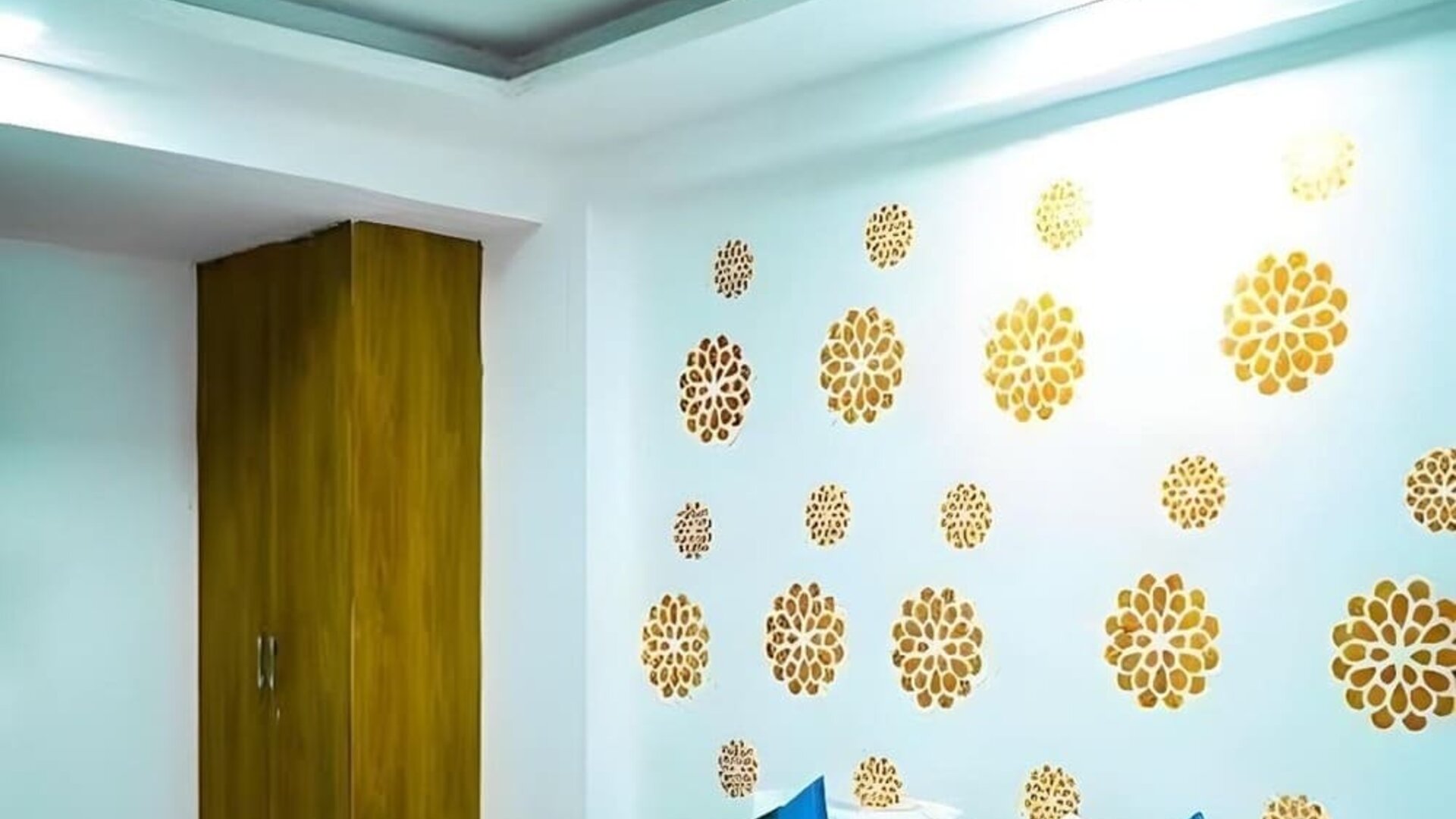 Guest room with a double bed, white bedding, teal and blue accent cushions, a matching bed runner, gold floral wall decorations, wooden wardrobe, and ceiling fan at Hotel Urban Abode.