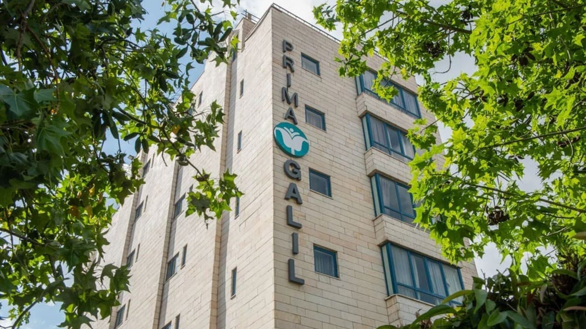 Exterior view of the hotel building surrounded by mature green trees at Prima Galil Tiberias.