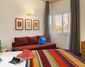 Guest bedroom with a colourful striped bedspread, red sofa, framed artwork and a flat-screen television at Prima Music Eilat.