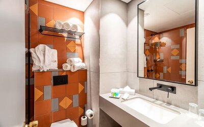 Bathroom with orange geometric accent tiles, a wall-mounted towel rack, bathrobes and a wide vanity mirror at Prima Music Eilat.