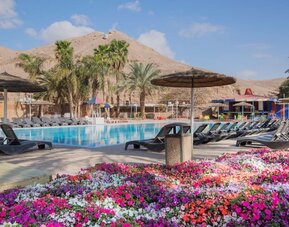 Outdoor swimming pool surrounded by sun loungers, parasols, palm trees and colourful flower beds at Prima Music Eilat.
