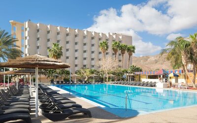 Outdoor swimming pool with rows of sun loungers, parasols and views of the hotel building and surrounding desert hills at Prima Music Eilat.
