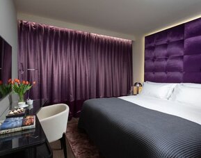 Guest room with king bed, purple upholstered headboard, matching curtains and work desk at Lady Stern Hotel.