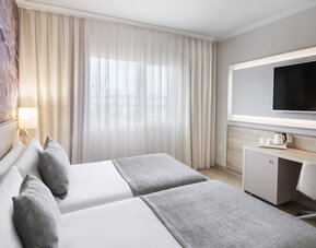 Twin bedroom with two single beds, grey runners, a desk, wall-mounted television and large curtained window at Hotel Sallés Ciutat del Prat.