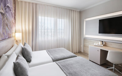 Twin bedroom with two single beds, grey runners, a desk, wall-mounted television and large curtained window at Hotel Sallés Ciutat del Prat.