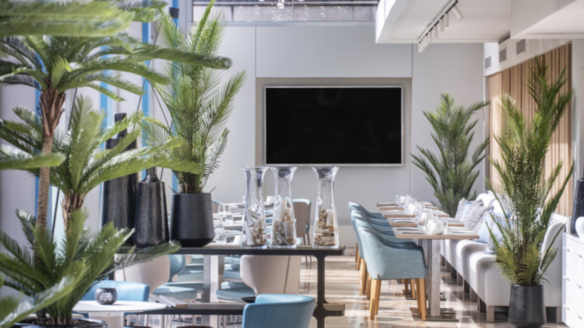 Bright dining lounge with blue and cream chairs, round tables, indoor palms and floor-to-ceiling windows at Hotel Sallés Ciutat del Prat.