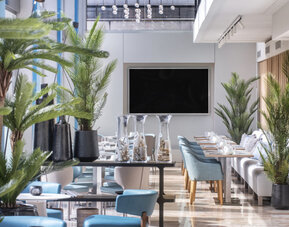 Bright dining lounge with blue and cream chairs, round tables, indoor palms and floor-to-ceiling windows at Hotel Sallés Ciutat del Prat.