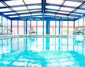 Indoor swimming pool beneath a glass roof, surrounded by tiled flooring and large windows at Hotel Sallés Ciutat del Prat.
