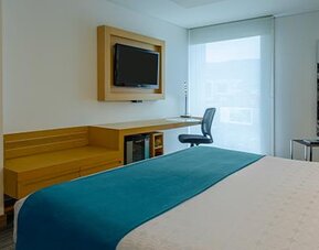 Guest room with white bedding, teal accent cushions, wooden desk and large window at Sonesta Hotel Bogotá.