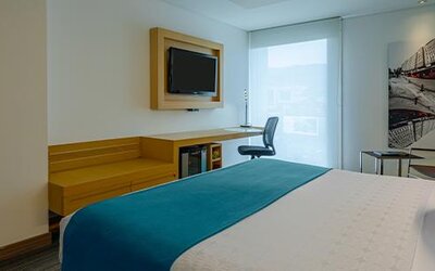 Guest room with white bedding, teal accent cushions, wooden desk and large window at Sonesta Hotel Bogotá.